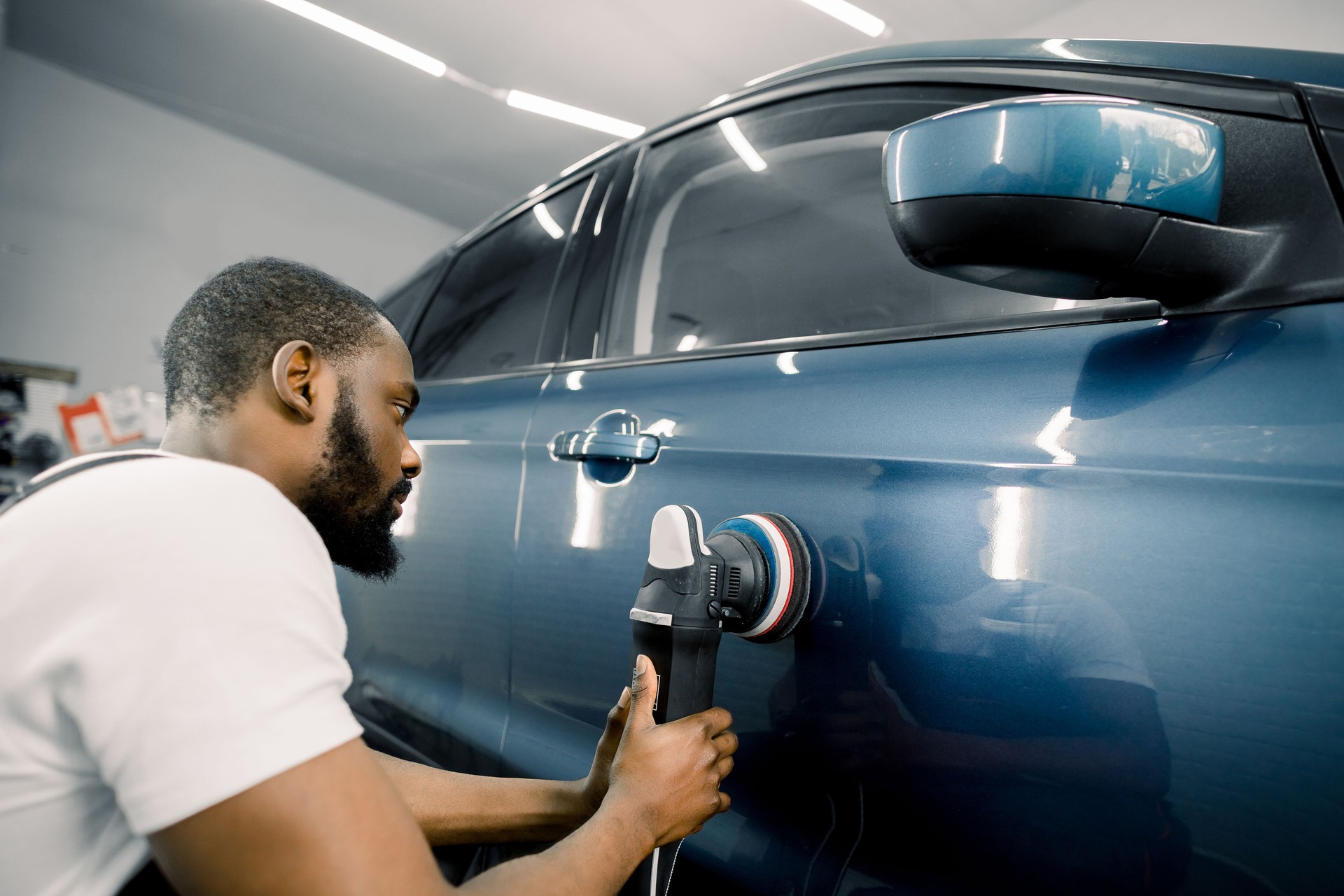 Young African man working at car detailing service, polishing blue car with polisher to eliminate contaminants from the surface of the car